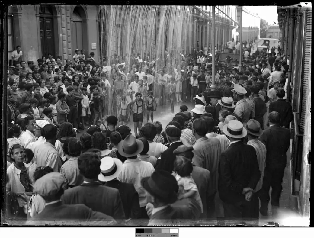 Foto histórica, 1940: una calle vista desde lo alto, con una cortina de agua que cae sobre la calzada, varios chicos debajo, algunos en ropa de baño, y una multitud apretada alrededor, con sombreros de paja en primer plano.