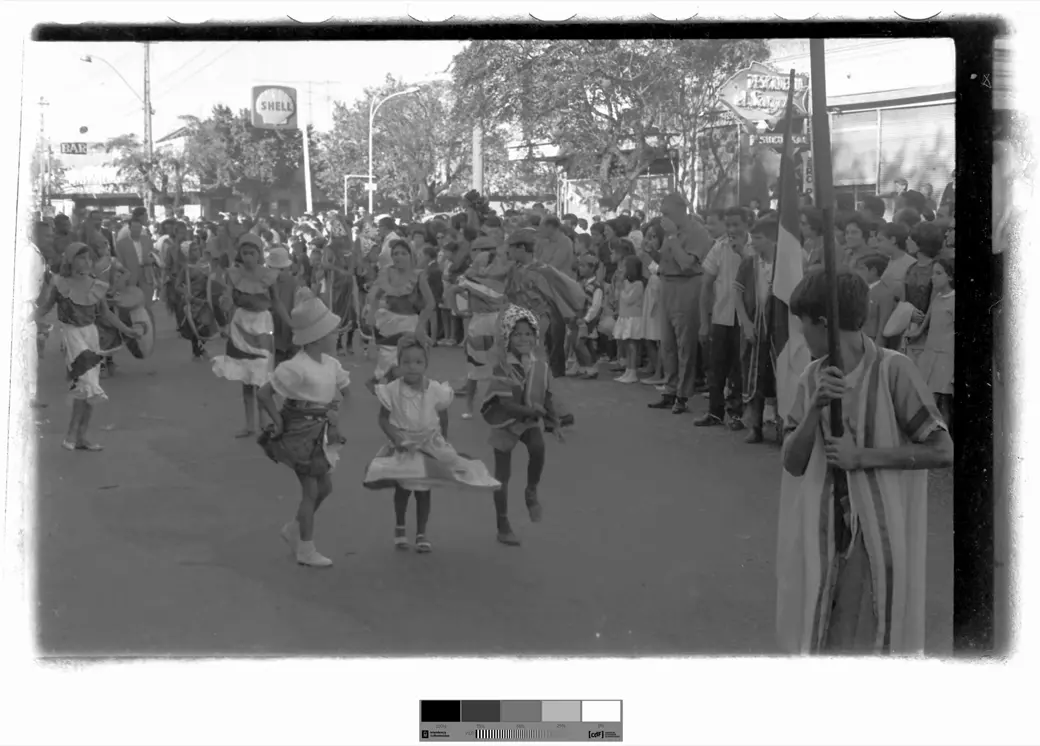 Foto histórica, 1966: tres chicos pequeños disfrazados bailan en medio de una calle asfaltada, con una fila de chicos con trajes de volados detrás, público de pie a la derecha y carteles de comercios al fondo.