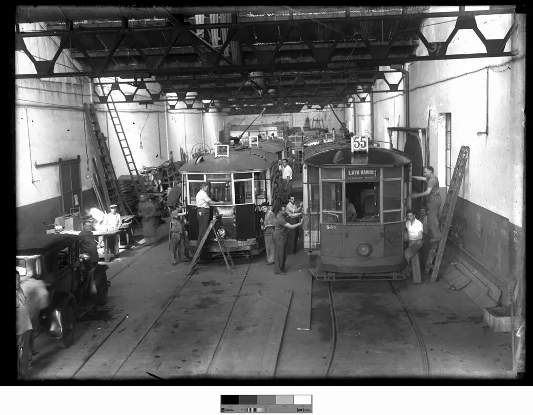 Un taller con techo de estructura metálica: tranvías sobre los rieles, dos en primer plano, uno con el cartel 55, rodeados de operarios y escaleras.