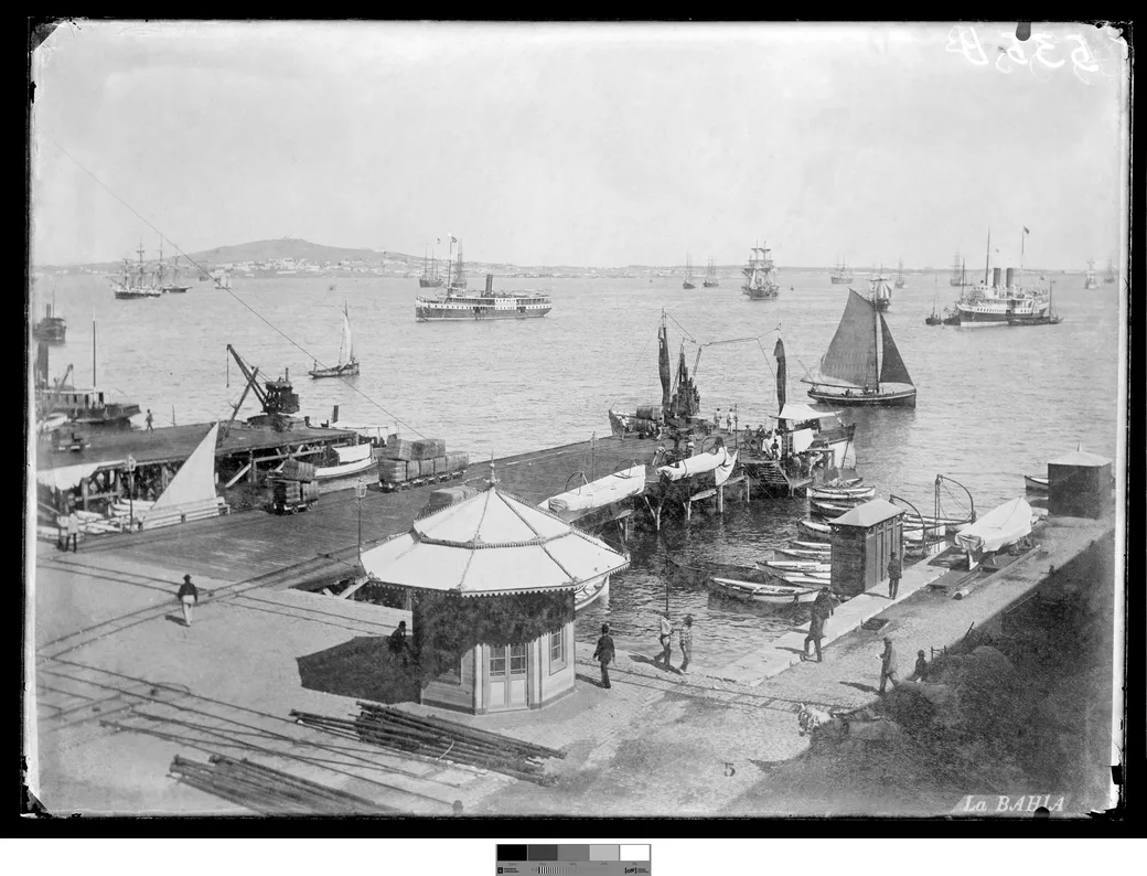 Foto histórica, década de 1890: la bahía de Montevideo con veleros y vapores fondeados, un pequeño muelle de madera en primer plano y el Cerro al fondo, sin obras portuarias.