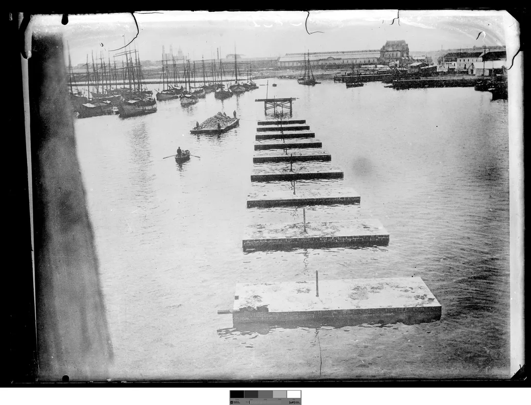 Foto histórica, 1901-1909: una fila de cajones de hormigón flotando alineados hacia la obra de la dársena 2, con un bote y una barcaza cargada cerca.