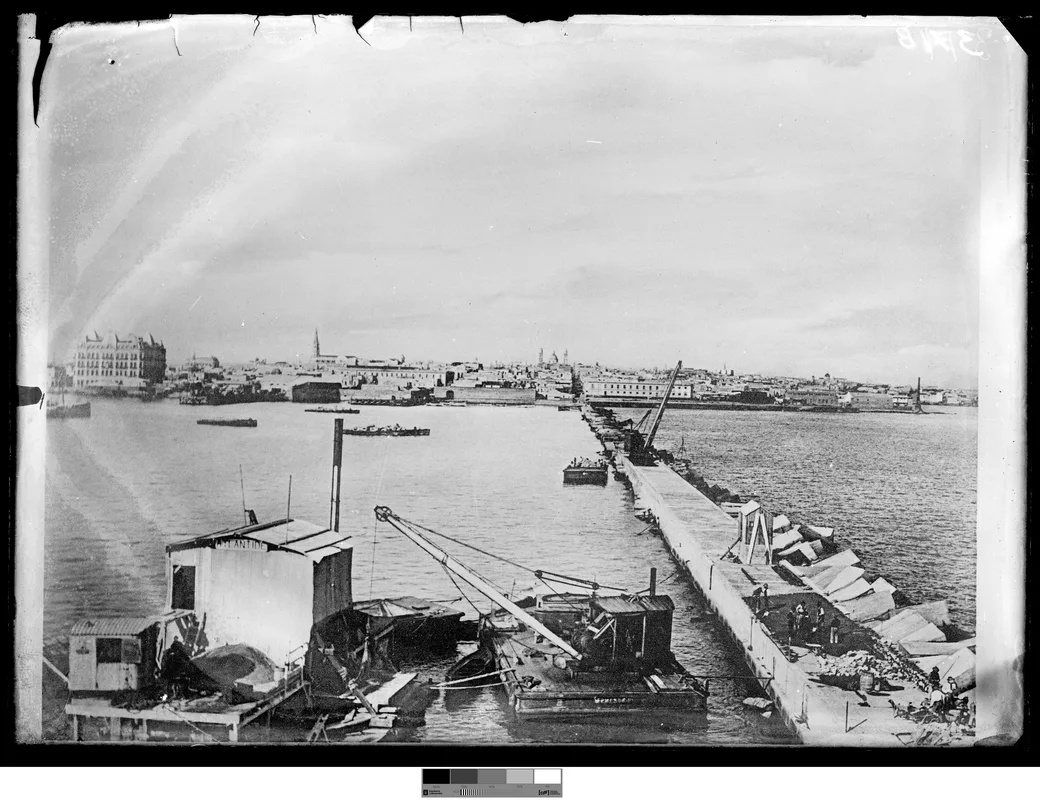 Foto histórica, 1901-1903: la escollera Sarandí en obra, con una plataforma flotante y grúas, bloques de piedra alineados y el Hotel Nacional y la Ciudad Vieja al horizonte.