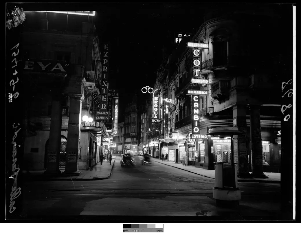 Foto histórica, 9 de mayo de 1951: Sarandí esquina Juncal de noche, con neón de casas de cambio y un auto en movimiento sobre la calzada.